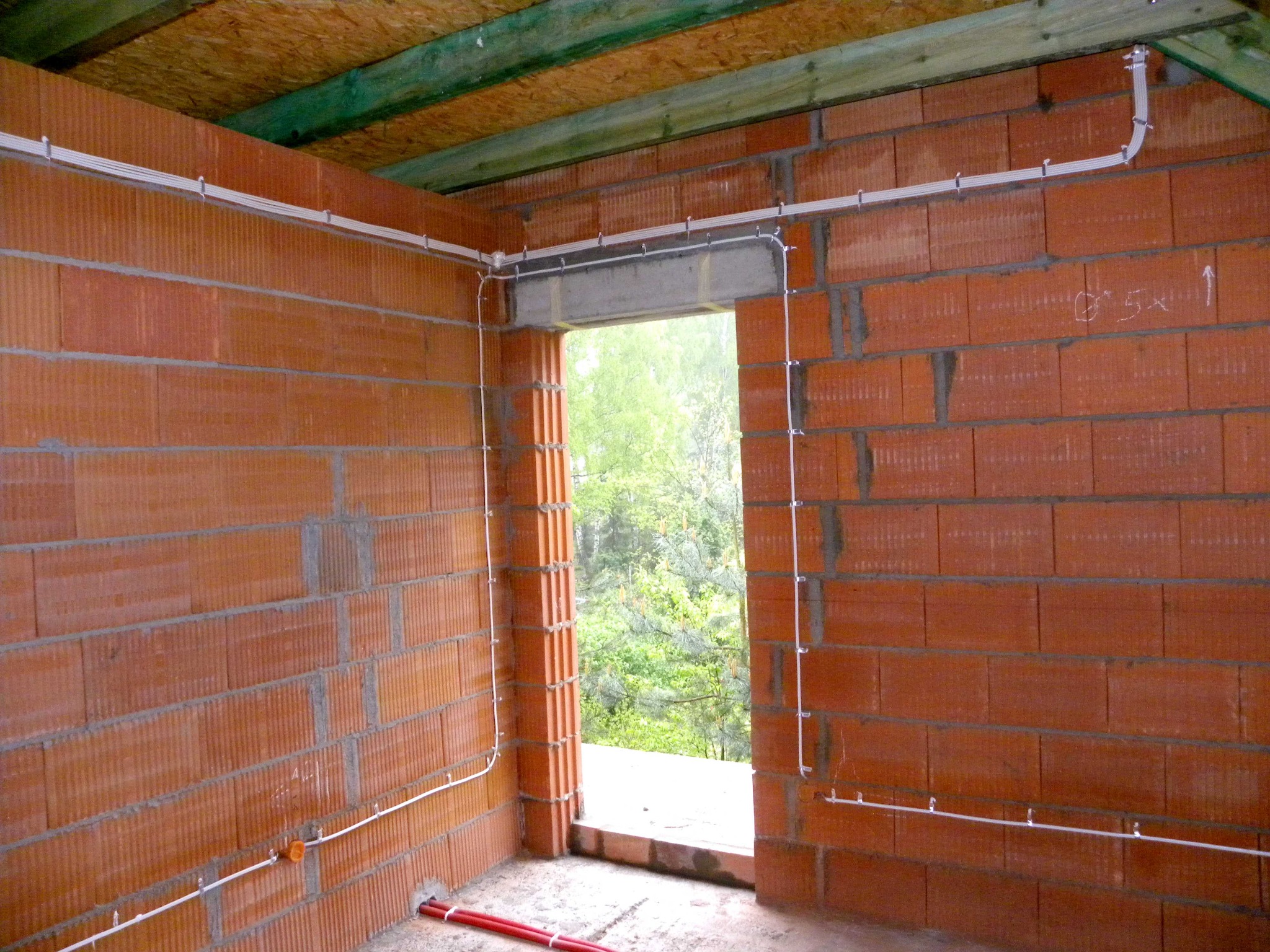 Wewnętrzne ściany z czerwonej cegły z widocznymi, prowadzonymi natynkowo białymi przewodami elektrycznymi mocowanymi uchwytami, widok na zewnątrz przez niezabezpieczony otwór okienny.