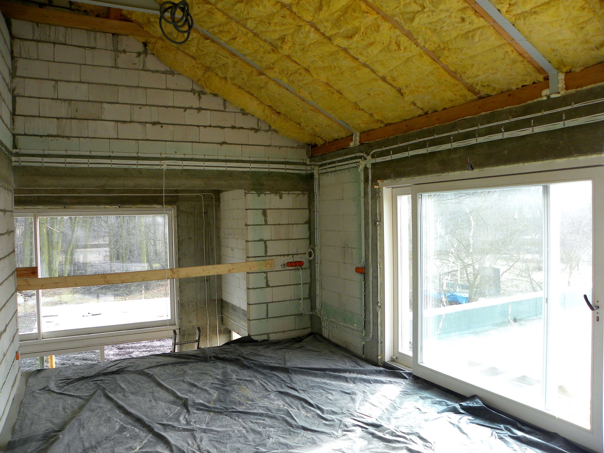 Wnętrze w trakcie prac wykończeniowych: surowy strop z izolacją, ściany z betonu komórkowego, rozprowadzone przewody elektryczne w peszlach, okna z widokiem na zewnątrz, podłoga zabezpieczona folią.