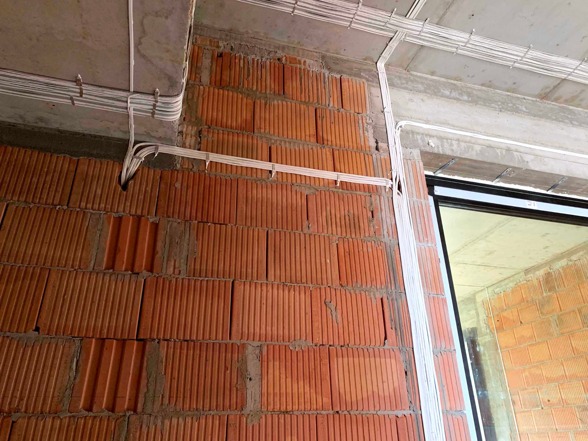 Instalacja elektryczna w surowym stanie: białe przewody prowadzone po ceglanej ścianie i betonowym suficie, mocowane za pomocą uchwytów, widoczne otwory w ścianie.