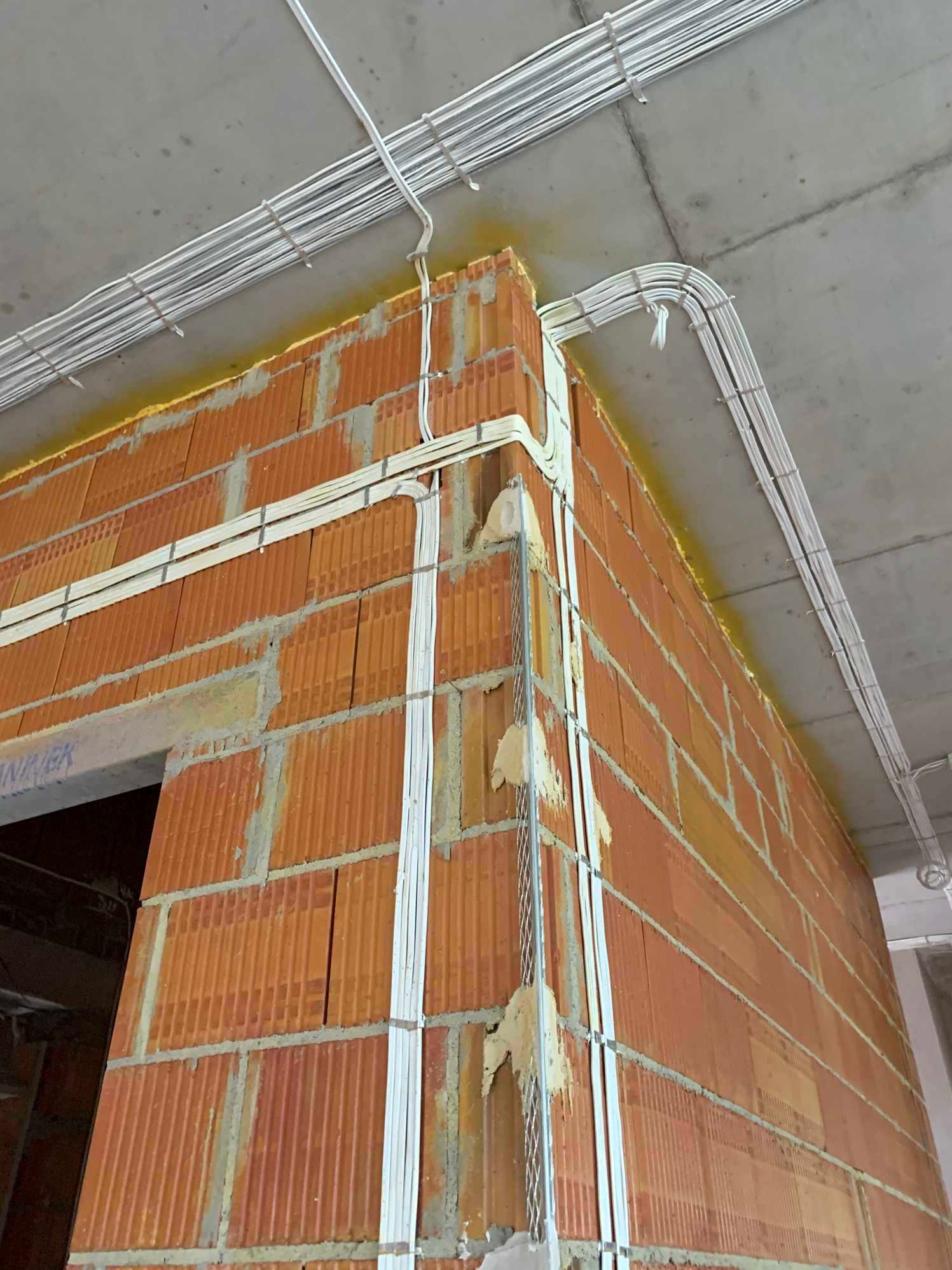 Świeżo położone przewody elektryczne w białych peszlach, prowadzone wzdłuż muru z czerwonej cegły i pod sufitem, zabezpieczone uchwytami. Widoczne strefy instalacyjne.