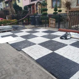 Częściowo ułożona kostka brukowa w szachownicę, czarno-biała, przed domem w Oławie; widoczne palety z kostką i narzędzia.