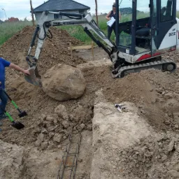 Mini koparka podnosi duży kamień podczas prac ziemnych przy budowie fundamentów, widoczny zbrojony wykop i pracownik z łopatą, w tle dom jednorodzinny.