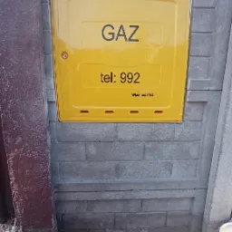 Ż&oacute;łta szafka gazowa z napisem GAZ i numerem telefonu 992, wbudowana w betonowe ogrodzenie, widoczna z poziomu gruntu.