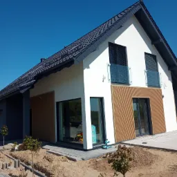 Nowoczesny dom jednorodzinny w Kościerzynie z białą elewacją, drewnianymi panelami i ciemną dachówką, w trakcie zagospodarowywania terenu wokół.