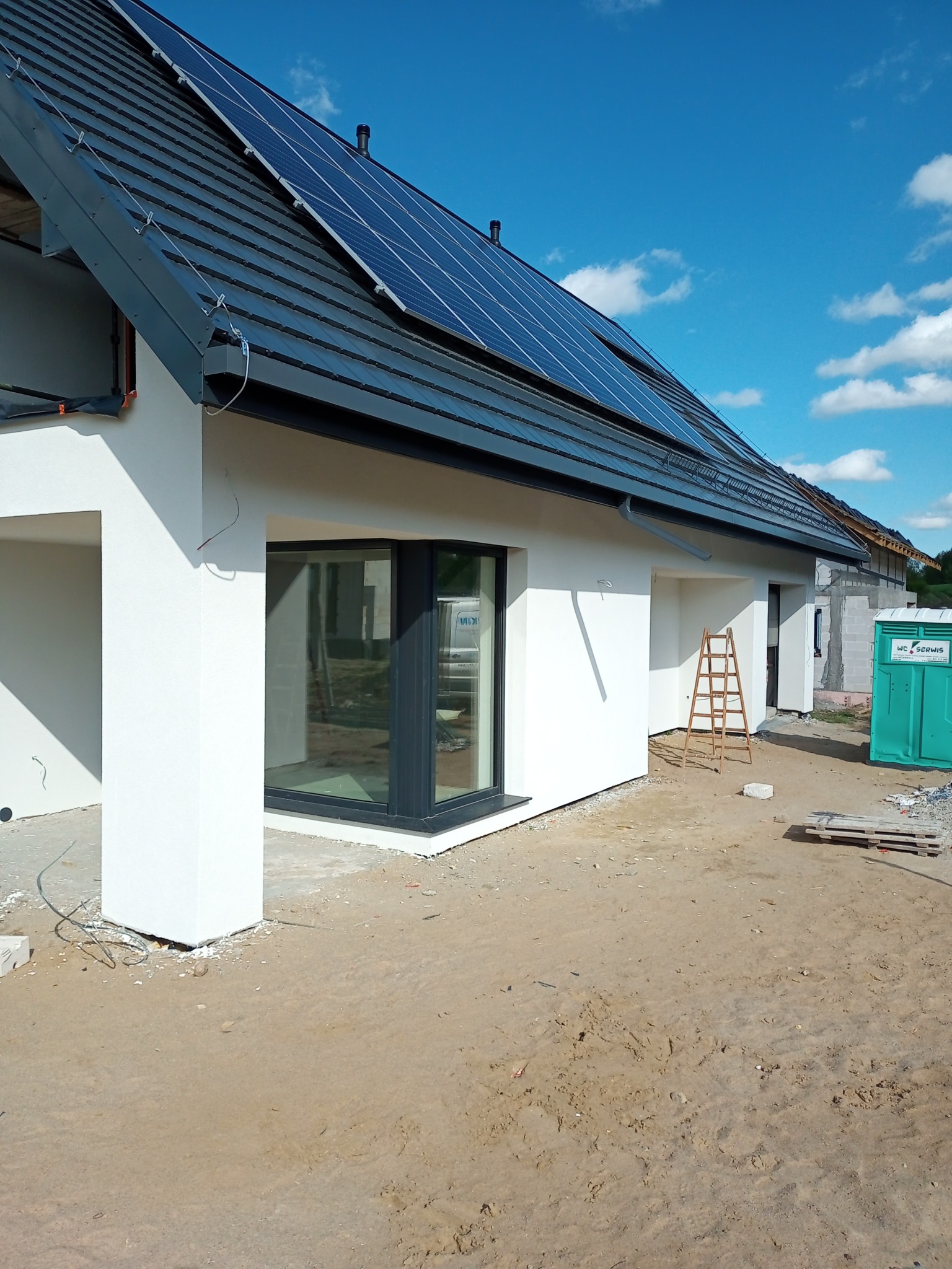 Niedokończony dom jednorodzinny z białymi ścianami, ciemną stolarką okienną i panelami słonecznymi na dachu, widoczna drabina i przenośna toaleta na zewnątrz.