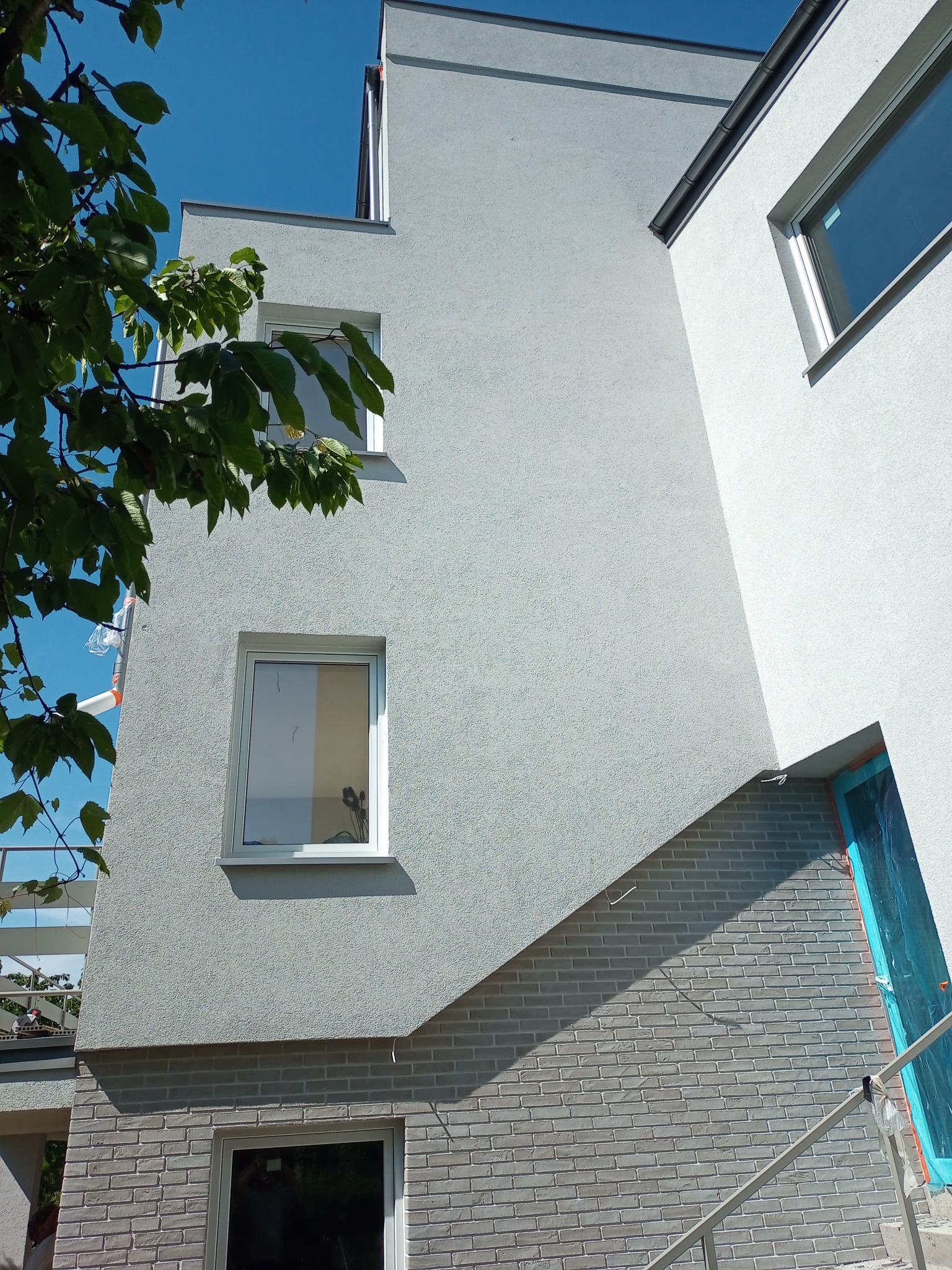 Fragment elewacji budynku z oknami, częściowo wykończonej szarym tynkiem strukturalnym i cegłą, zabezpieczone folią okno w narożniku, widok z dołu w słoneczny dzień.