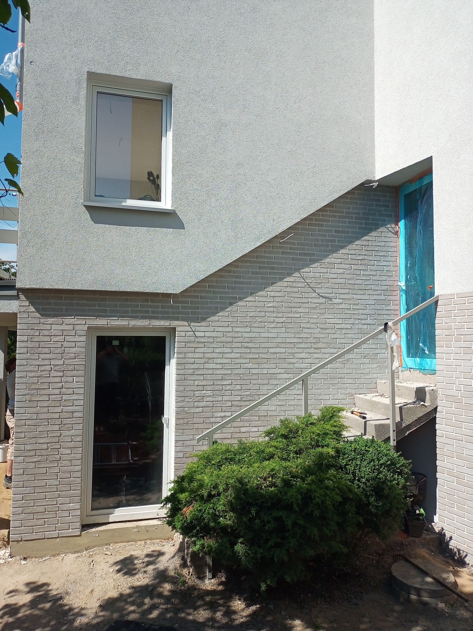 Fragment elewacji budynku z oknami i schodami zewnętrznymi; dolna część ściany wykończona szarą cegłą klinkierową, górna pokryta jasnoszarym tynkiem strukturalnym; drzwi wejściowe zabezpieczone...