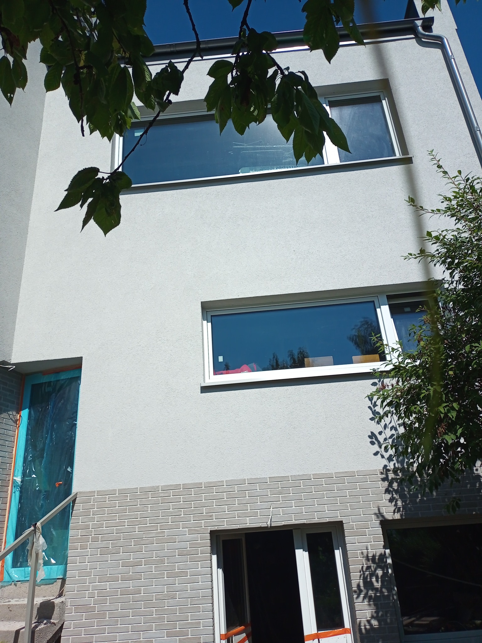 Szara elewacja domu z widocznym fragmentem okładziny z cegły, okna z białymi ramami, drzwi zabezpieczone folią ochronną i fragment rusztowania.