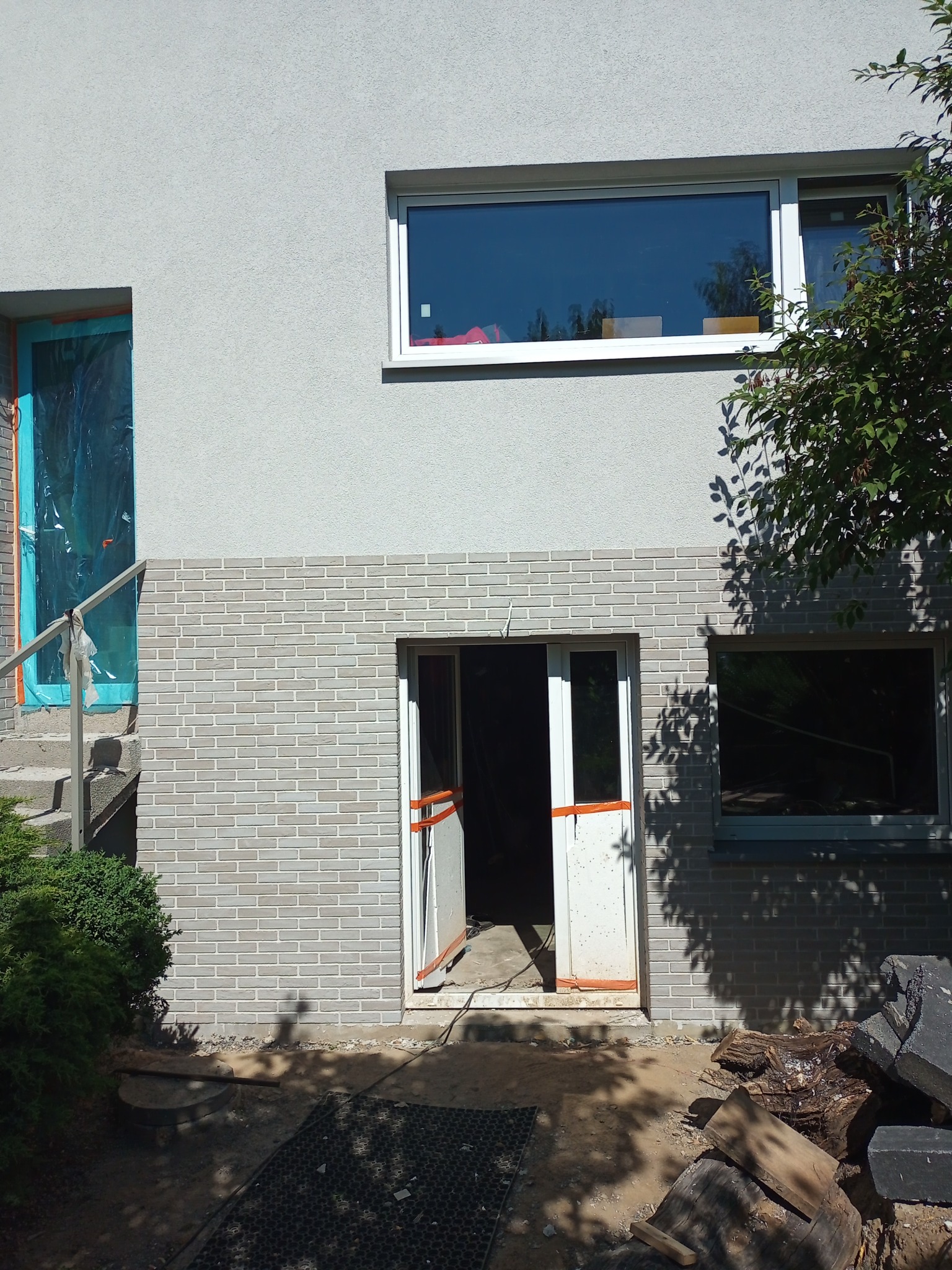 Dwukondygnacyjny budynek z szarą elewacją i fragmentem ściany wykończonym cegłą klinkierową, okno zabezpieczone folią ochronną, otwarte drzwi wejściowe z pomarańczowymi zabezpieczeniami, fragment...