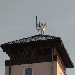 Srebrna syrena alarmowa z anteną zamontowana na dachu budynku z drewnianą konstrukcją i oknami w pochmurny dzień.