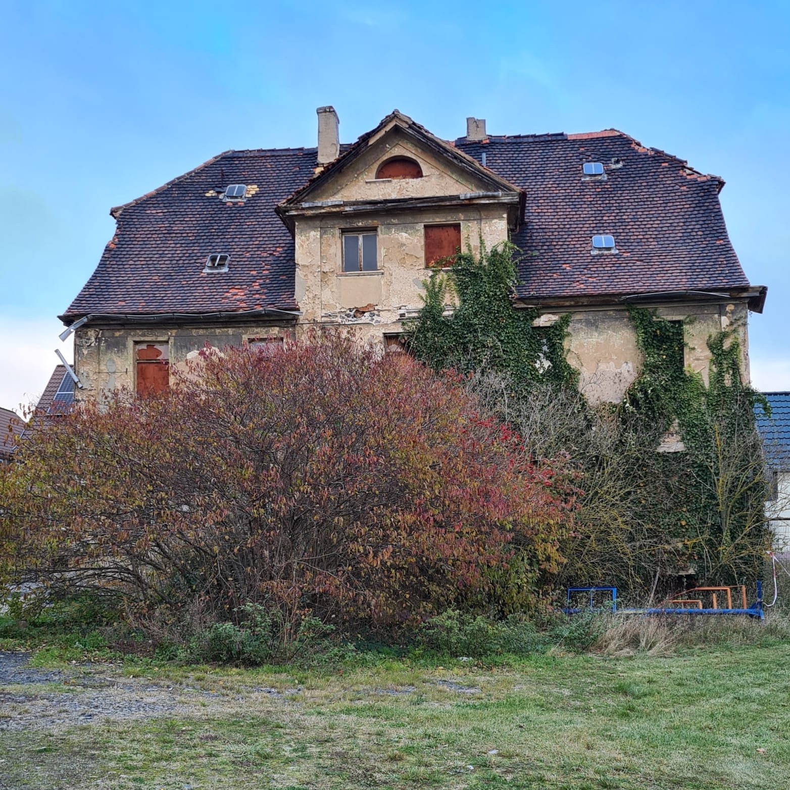 Zrujnowany, dwupiętrowy budynek z ceglanym dachem, częściowo porośnięty bluszczem, z zasłoniętymi oknami i zniszczoną fasadą, widoczny zza krzewów o czerwonych liściach.