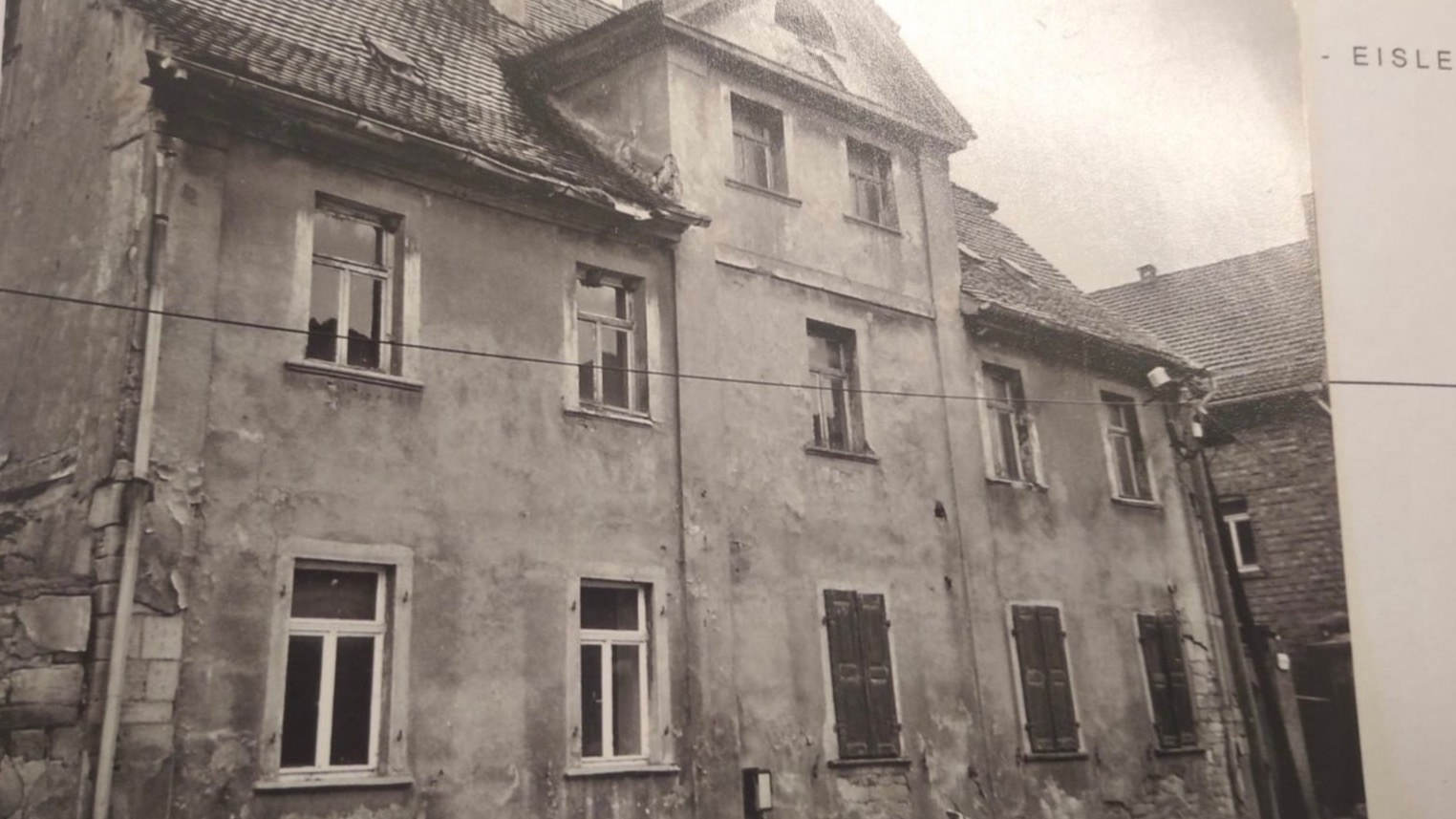 Czarno-białe zdjęcie elewacji starej, zaniedbanej kamienicy z widocznymi uszkodzeniami tynku, niektórymi oknami z okiennicami i fragmentem dachu sąsiedniego budynku.