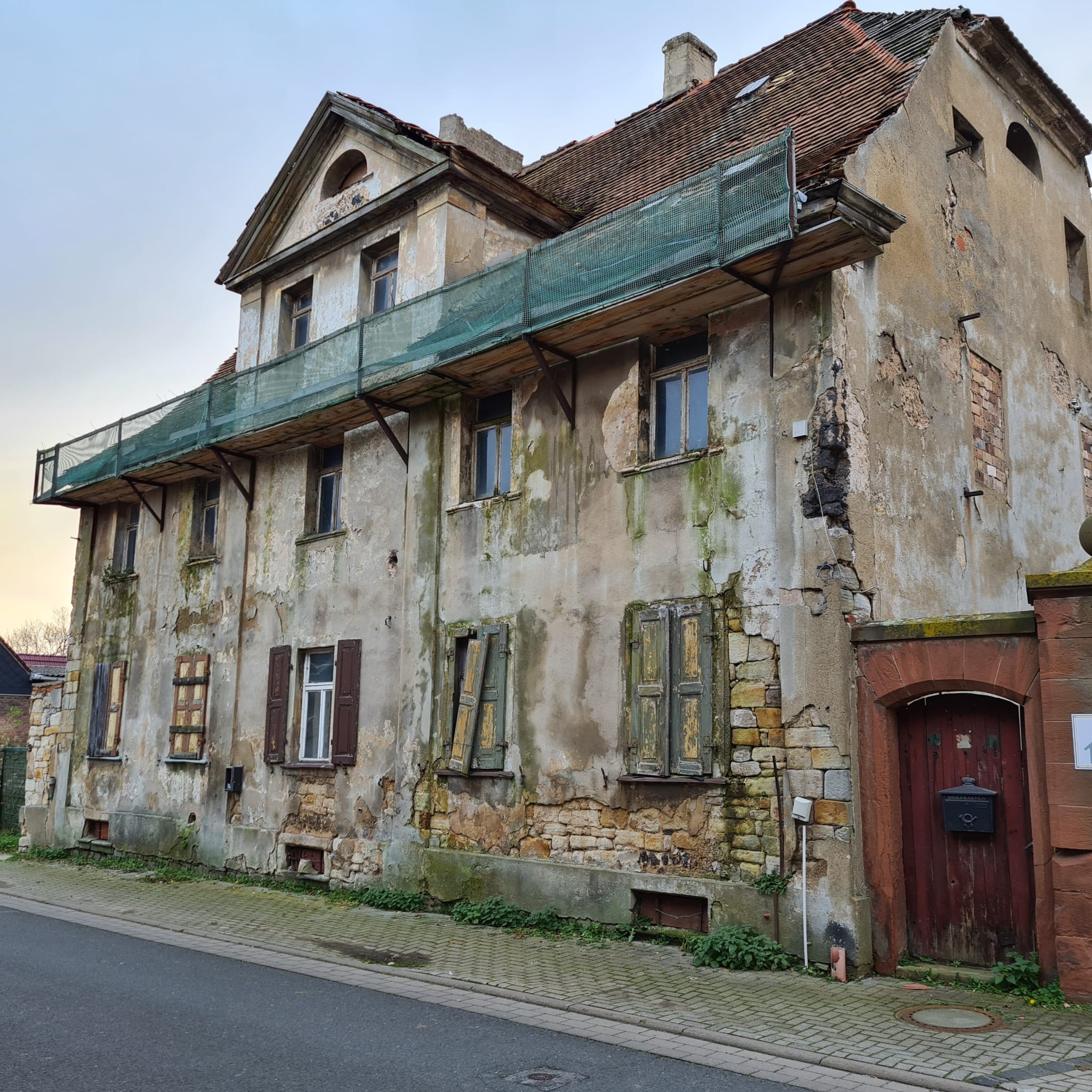 Zniszczona elewacja starej kamienicy z fragmentami rusztowania i siatki zabezpieczającej, widoczne ubytki tynku i odsłonięta cegła, częściowo zasłonięte okna z drewnianymi okiennicami.