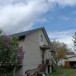 Dom jednorodzinny z szarym dachem z blachodachówki, częściowo ocieplony, z widocznym rusztowaniem i narzędziami dekarskimi, w otoczeniu kwitnących bzów i garażu z samochodem.