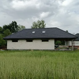 Niedokończony budynek gospodarczy z czarnym dachem z dachówki ceramicznej i dwoma oknami dachowymi, widoczne otwory okienne w ścianach z betonu komórkowego, w tle zieleń i pochmurne niebo.