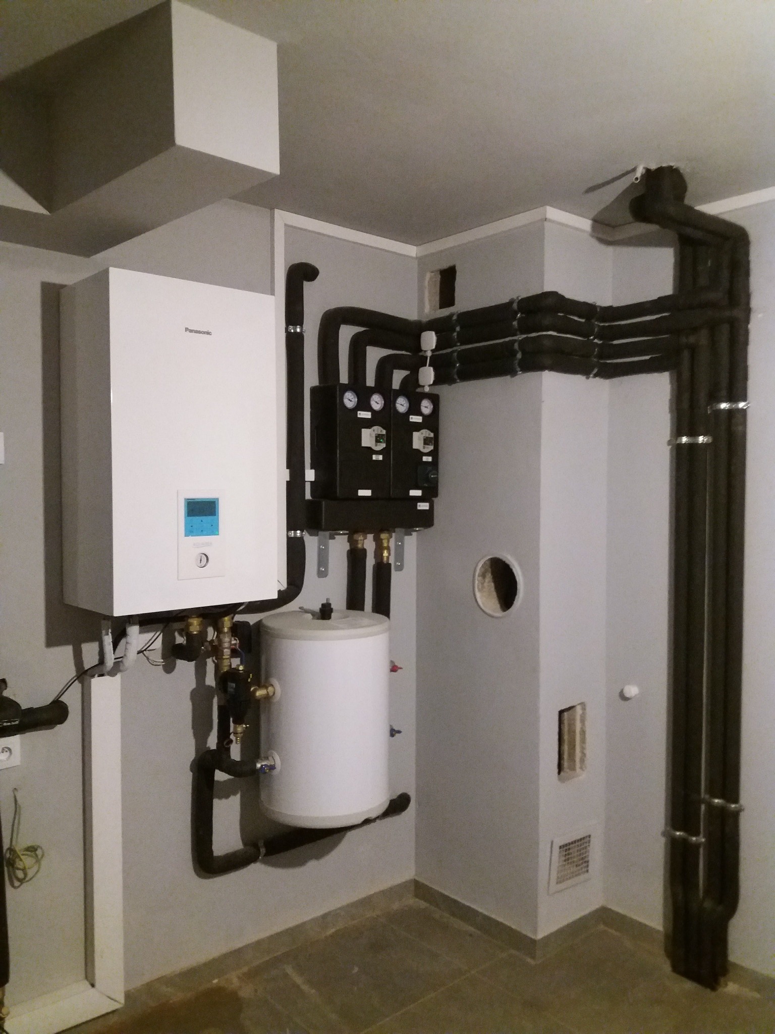 Biała jednostka wewnętrzna pompy ciepła Panasonic, zbiornik akumulacyjny i czarna szafka sterownicza z manometrami, zainstalowane na szarej ścianie w pomieszczeniu technicznym. Izolowane rury...