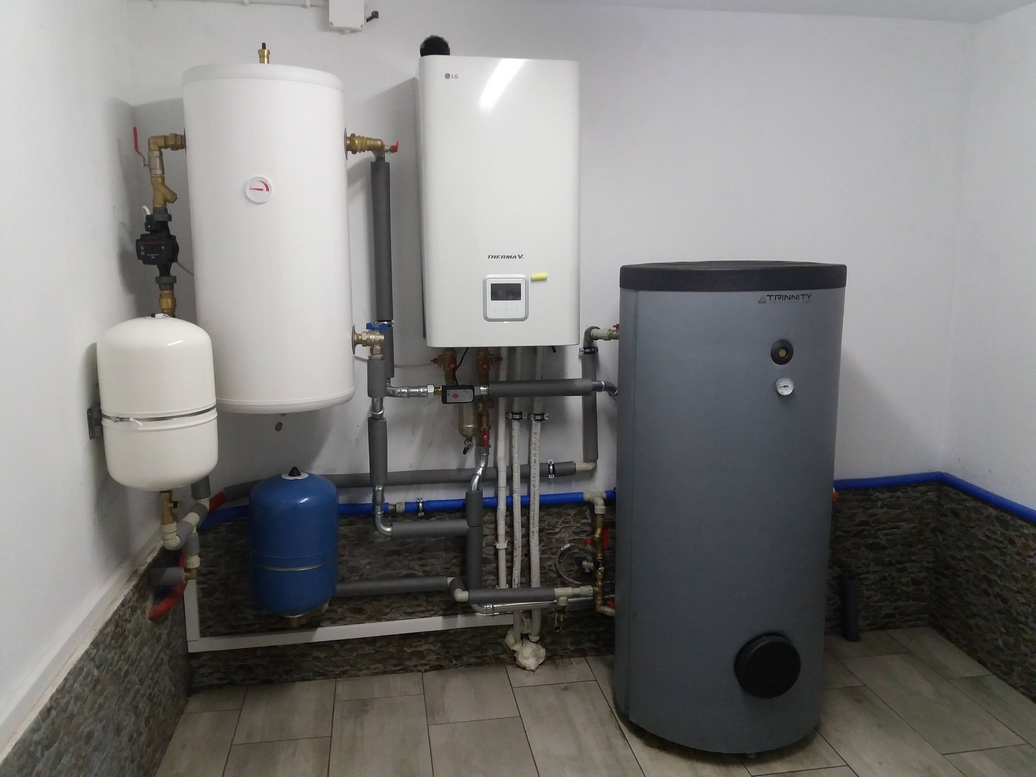 Zestaw urządzeń grzewczych w kotłowni: biały zasobnik ciepłej wody z widocznym manometrem, wiszący biały piec gazowy LG Therma V z wyświetlaczem oraz szary zbiornik buforowy Trinitty, połączone...