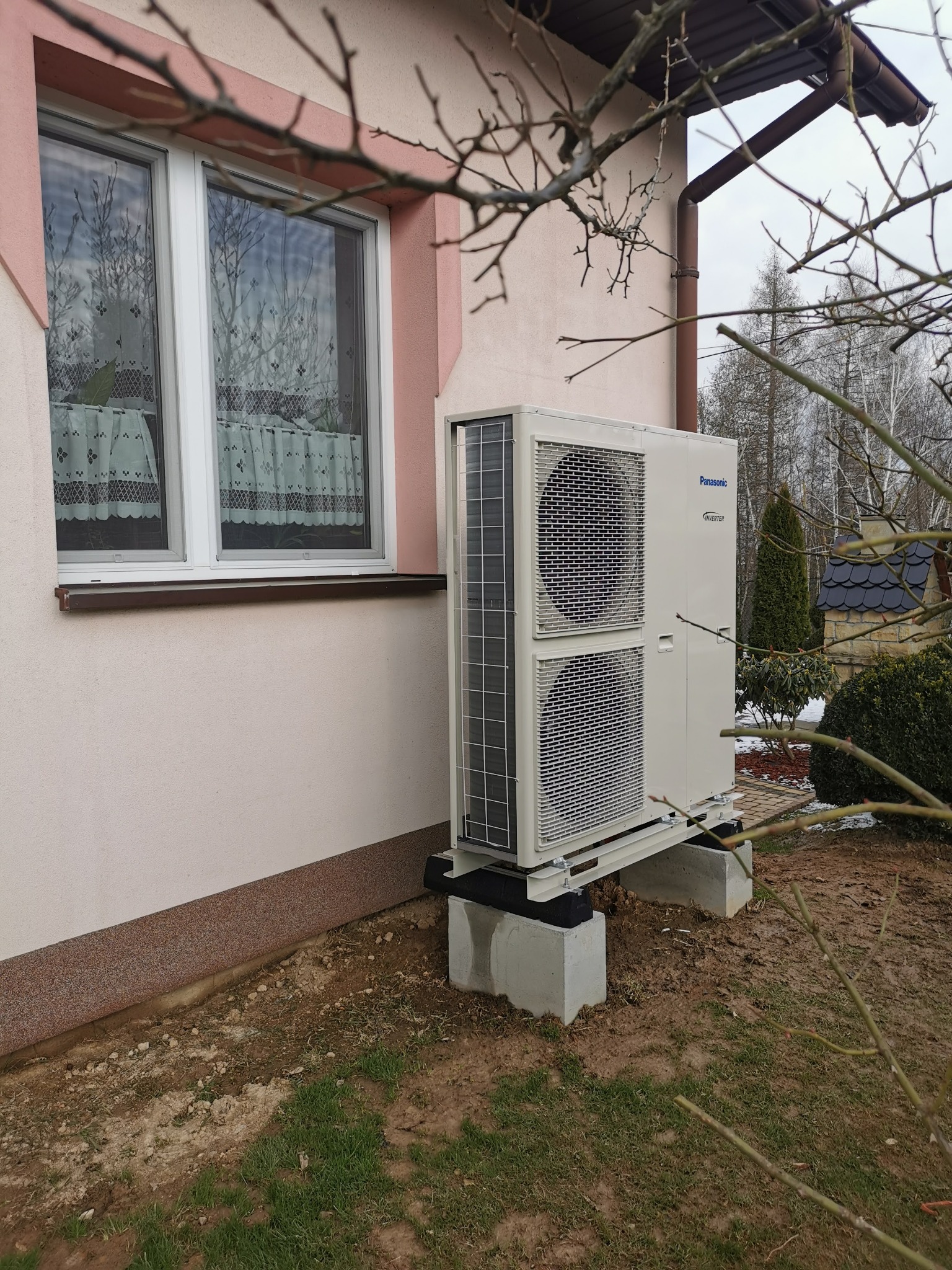 Biała, zewnętrzna jednostka klimatyzacji Panasonic Inverter zamontowana na betonowych podstawach przy ścianie domu z oknem z firankami, widoczne gałęzie drzew.