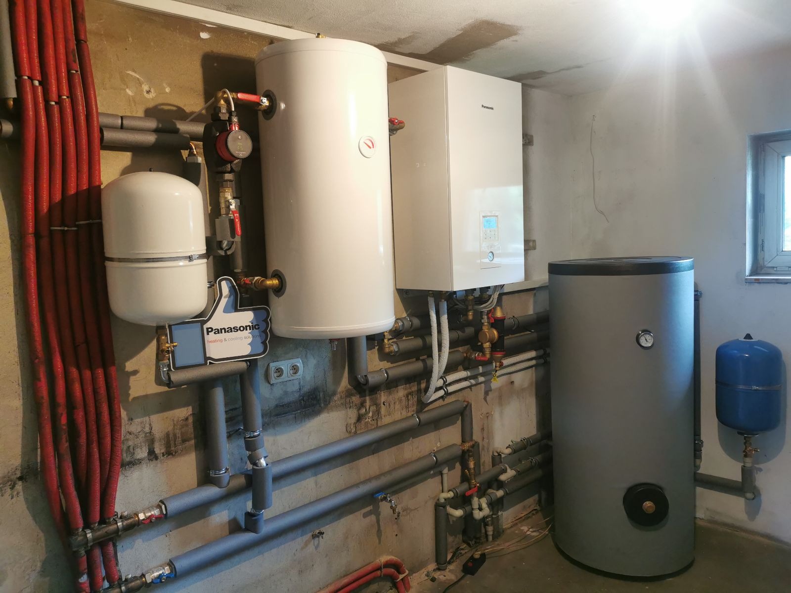 Instalacja grzewcza w piwnicy: biały zasobnik ciepłej wody, szary zbiornik buforowy, biały moduł pompy ciepła Panasonic, rury w izolacji, znak 'kciuk w górę' Panasonic na rurach.