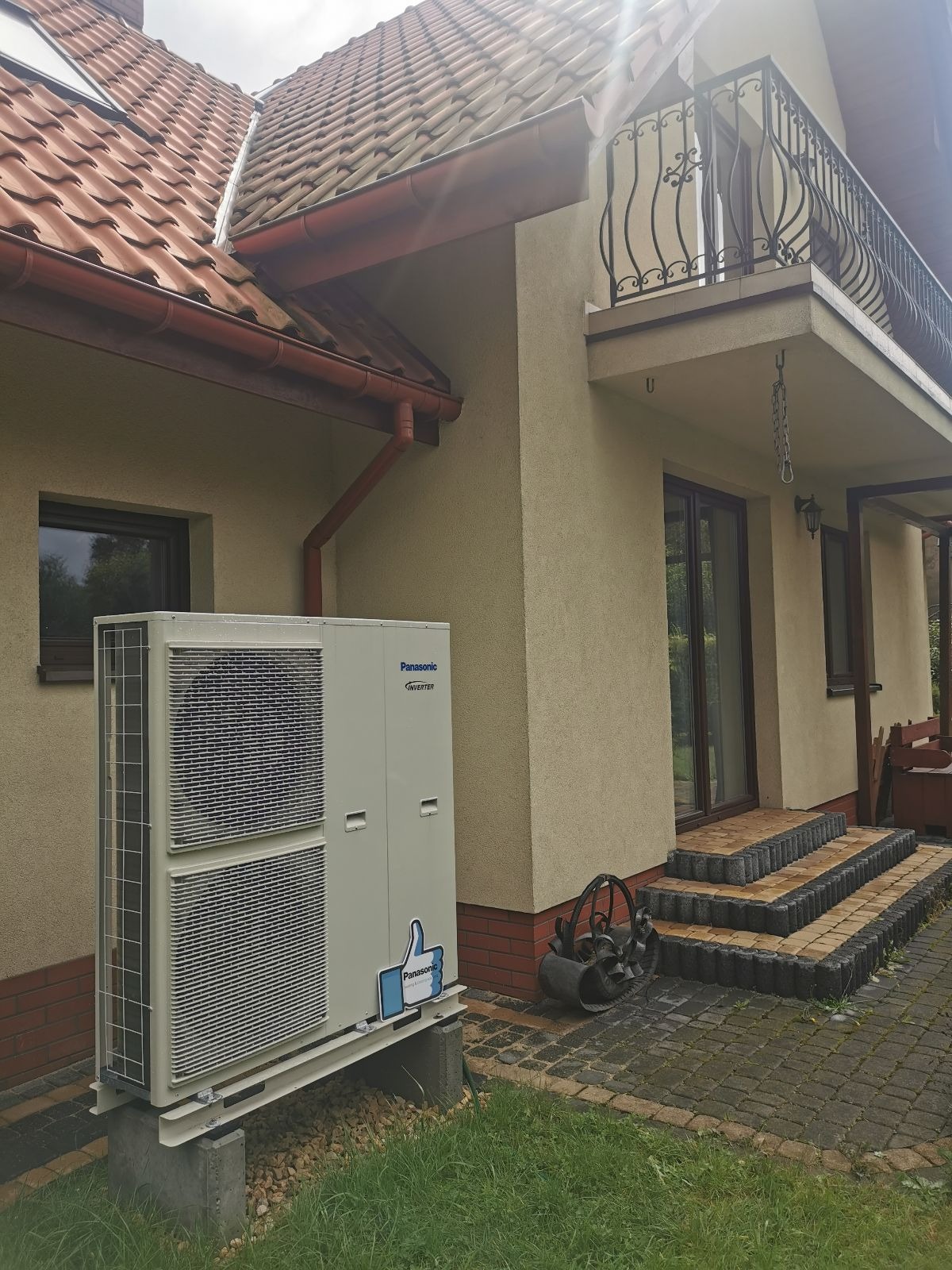 Zewnętrzna jednostka pompy ciepła Panasonic Inverter zamontowana na betonowych blokach przy ścianie domu z czerwoną dachówką, obok wejścia z kamiennymi schodami i ozdobnym balkonem.