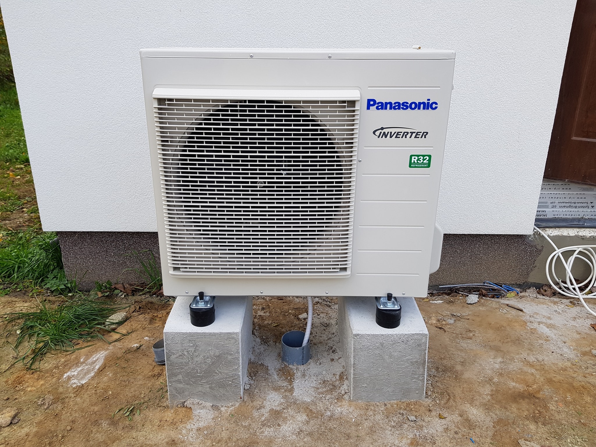 Zewnętrzna jednostka klimatyzacji Panasonic Inverter R32, zamontowana na betonowych bloczkach z regulowanymi nóżkami, przy białej ścianie budynku. Widoczne przewody i rury przyłączeniowe.