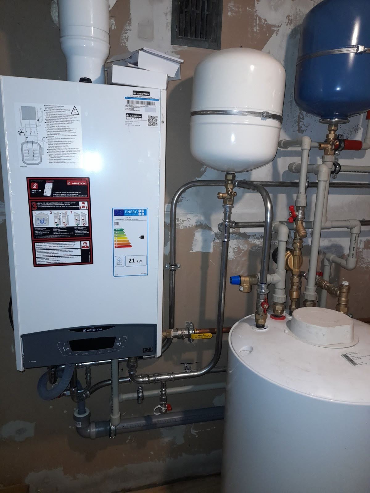 Nowoczesny piec gazowy Ariston 21kW z podłączonymi zbiornikami wyrównawczymi i instalacją hydrauliczną na tle ściany z widocznymi śladami po remoncie.