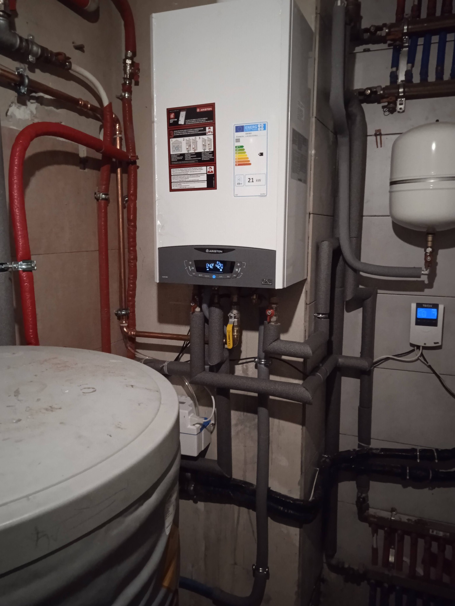 Nowoczesny piec gazowy Ariston z podłączonymi rurami w izolacji, widoczny zbiornik akumulacyjny i panel sterowania, instalacja w pomieszczeniu technicznym.