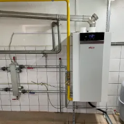 Biały piec gazowy Elco z podłączonymi rurami gazowymi (żółta) i wodnymi (srebrne) na tle kafelkowanej ściany, widoczny zbiornik wyrównawczy.