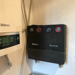 JM - Biały piec gazowy Buderus z podłączonymi dwoma czarnymi modułami pomiarowymi z tarczami w kolorze czerwonym i niebieskim, zamontowany na białej ścianie w kotłowni.