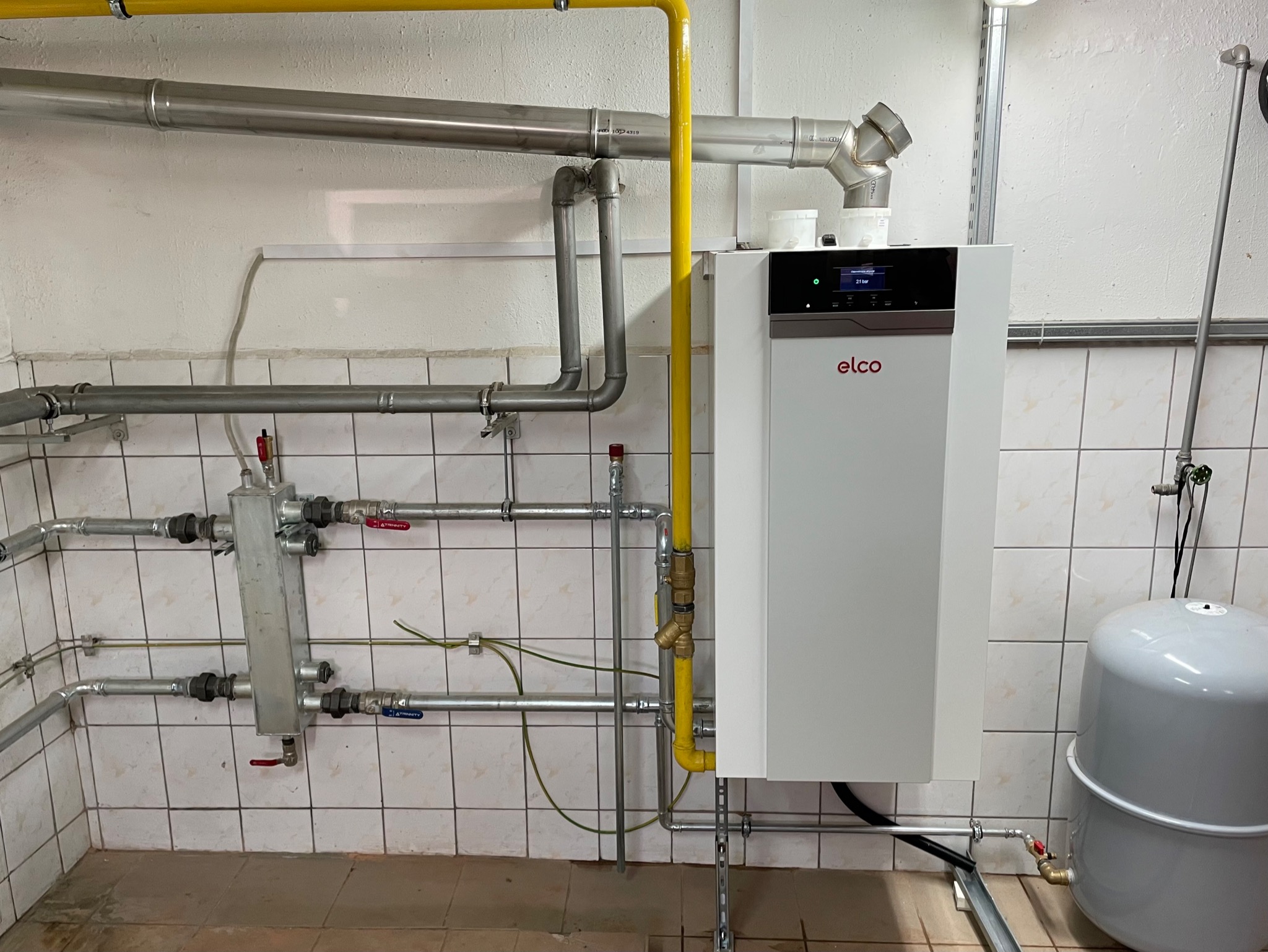 Biały piec gazowy Elco z podłączonymi rurami gazowymi (żółta) i wodnymi (srebrne) na tle kafelkowanej ściany, widoczny zbiornik wyrównawczy.