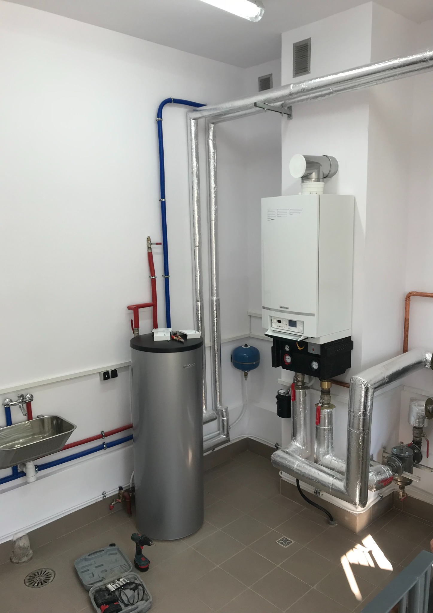 Nowoczesna kotłownia: biały piec gazowy, srebrny zasobnik ciepłej wody, izolowane rury, narzędzia w kuferku na podłodze. Czyste, białe ściany i płytki podłogowe.