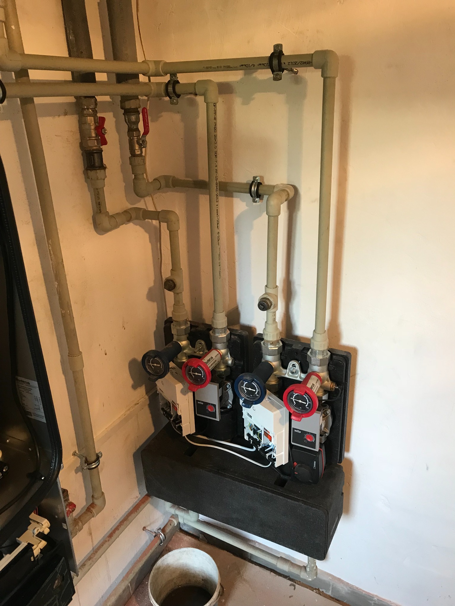 Rozbudowana instalacja grzewcza z rurami z tworzywa, zaworami z czerwonymi pokrętłami i dwoma czarnymi blokami z manometrami Wilo, widoczna również część kotła Buderus i wiadro na podłodze.