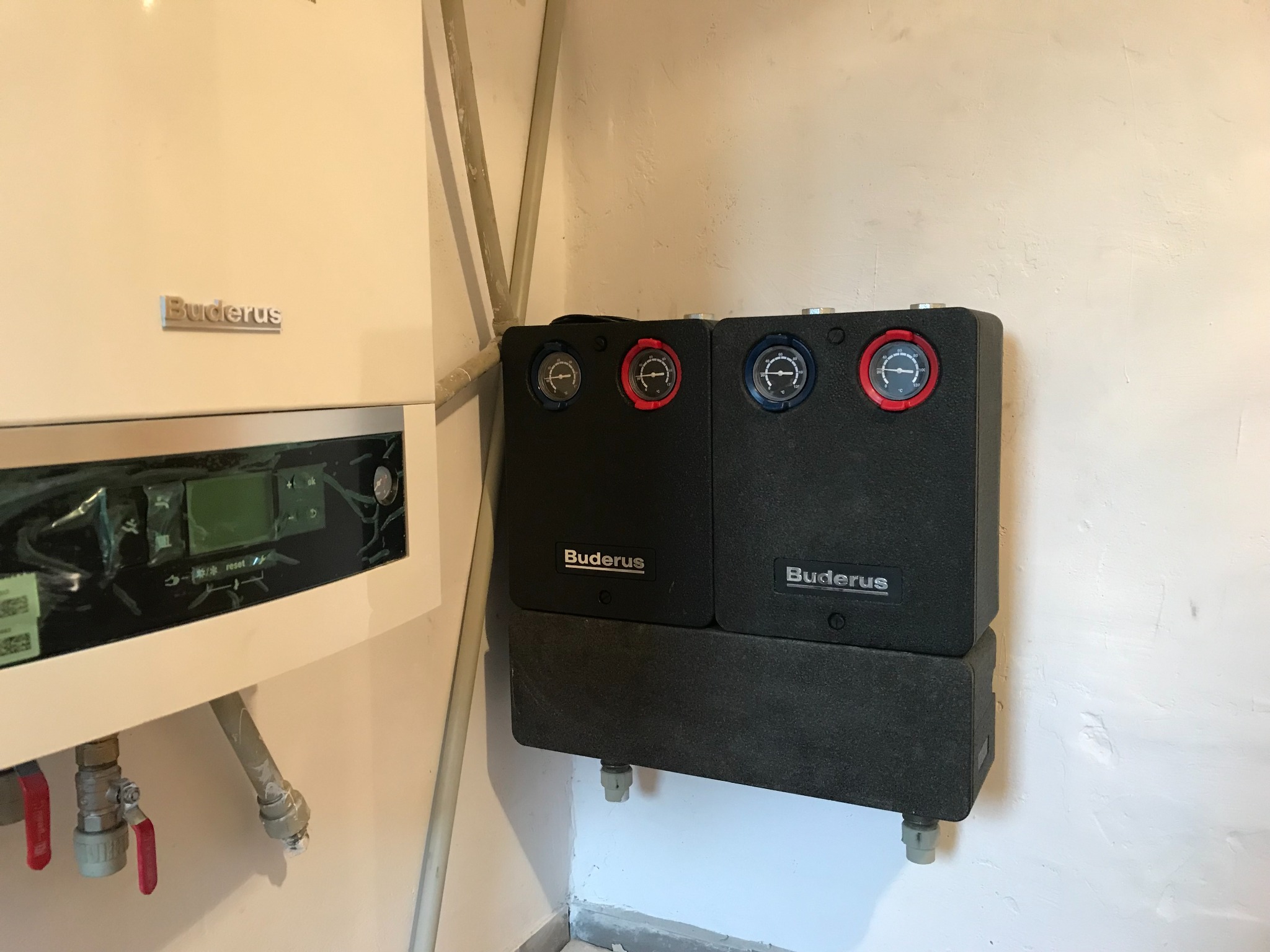 Biały piec gazowy Buderus z podłączonymi dwoma czarnymi modułami pomiarowymi z tarczami w kolorze czerwonym i niebieskim, zamontowany na białej ścianie w kotłowni.