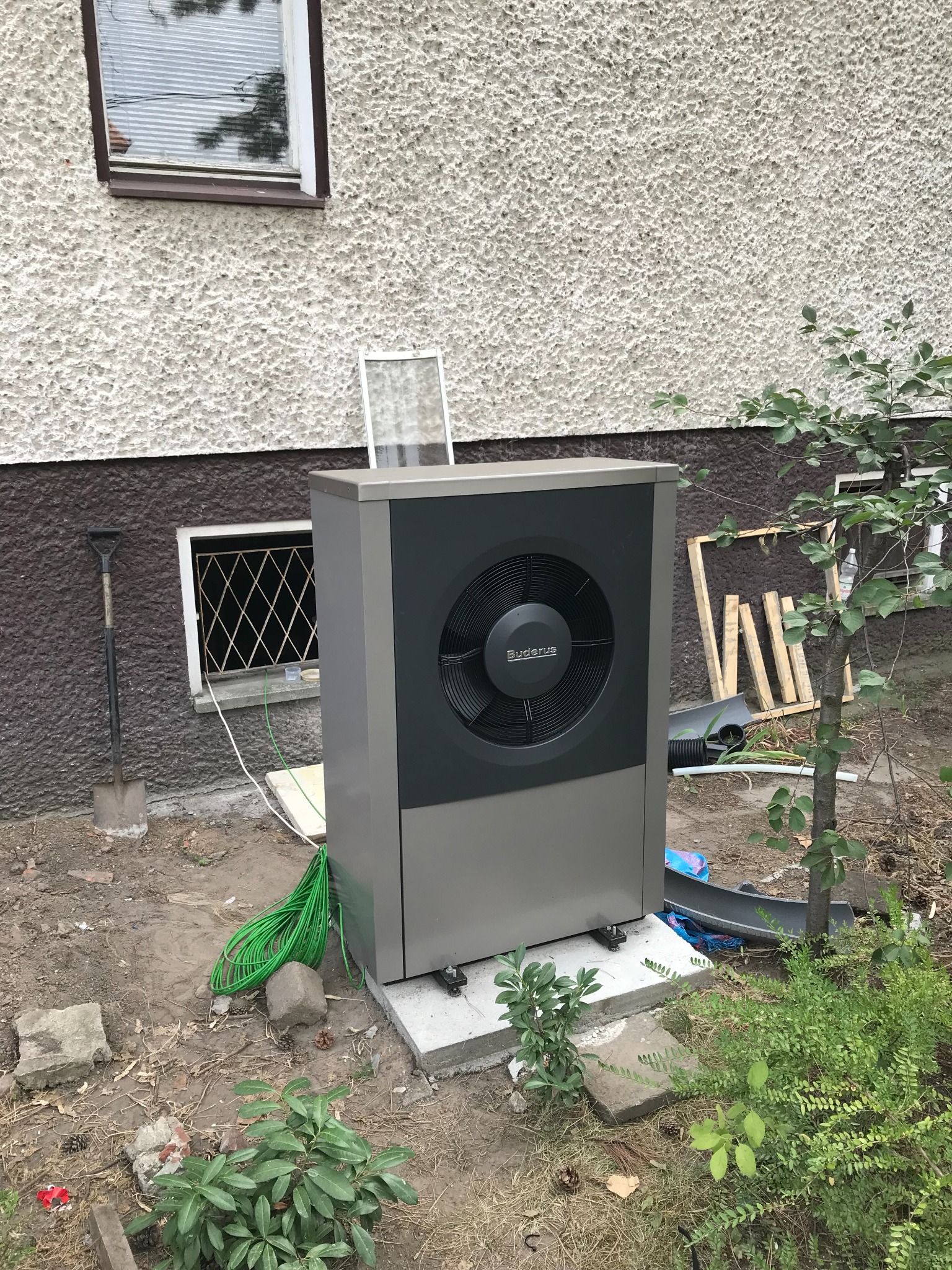Szaro-czarna pompa ciepła Buderus ustawiona na betonowej podstawie przy ścianie budynku z widoczną łopatą, zielonym kablem i roślinnością.
