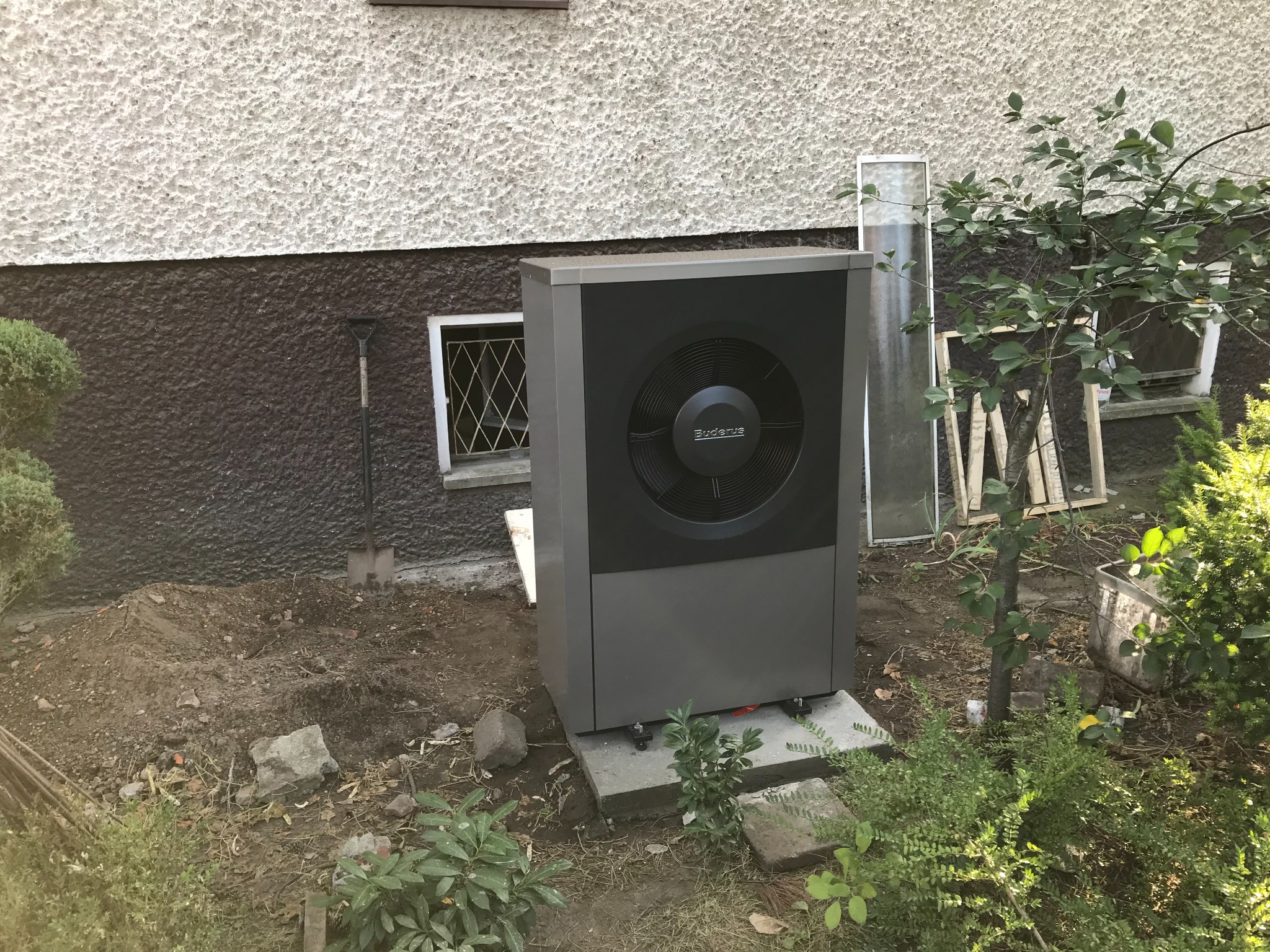 Szara pompa ciepła Buderus zainstalowana na betonowym fundamencie przy ścianie budynku z brązowo-białą elewacją, obok okna z kratą i łopaty wbitej w ziemię.