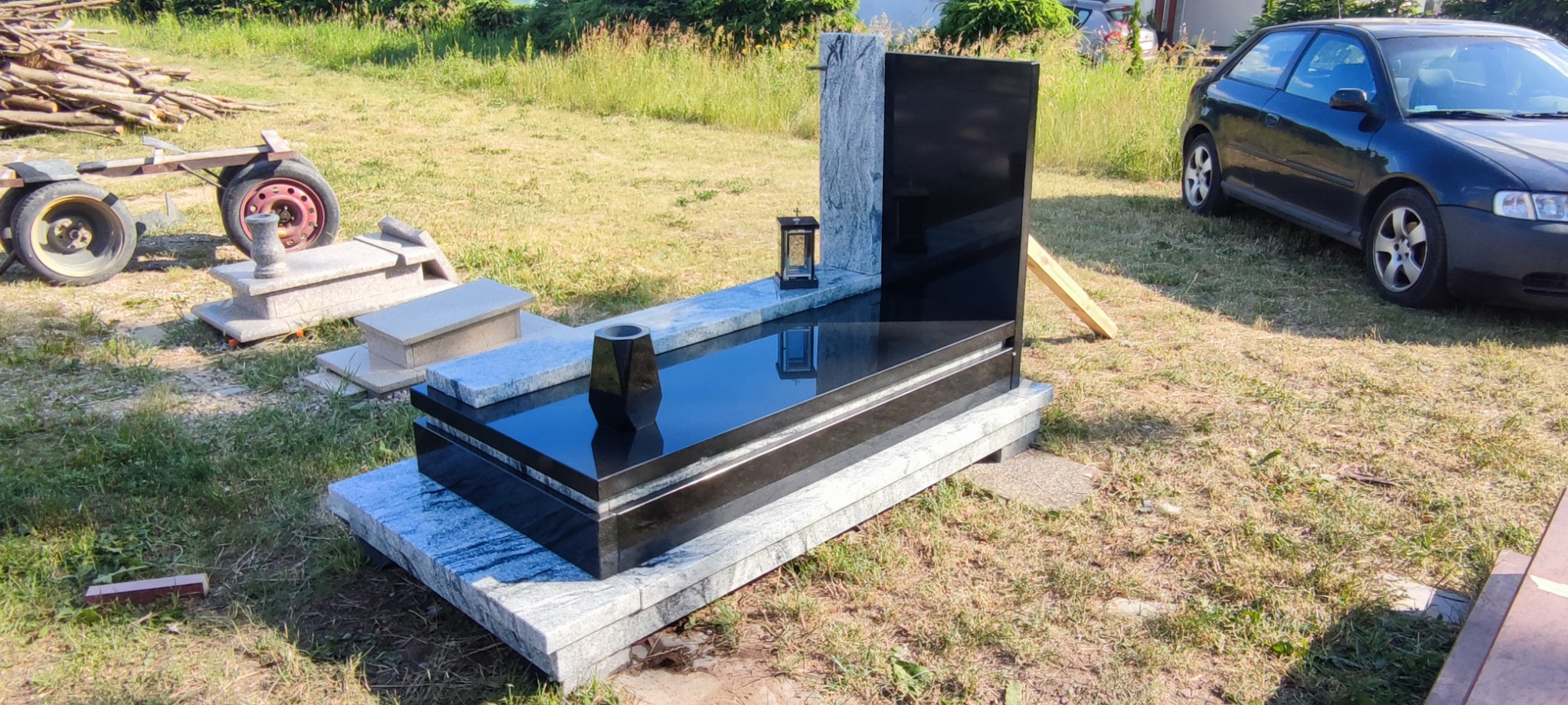 Nowoczesny, czarno-szary pomnik z wazonem i lampionem na trawiastym terenie, w tle zaparkowany samochód i przyczepka z drewnem.