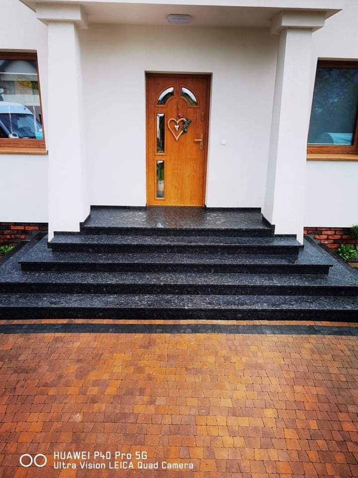 Zewnętrzne schody wejściowe wyłożone czarnym, nakrapianym kamieniem, prowadzące do drewnianych drzwi z sercem, na tle białego budynku z kolumnami i kostki brukowej.