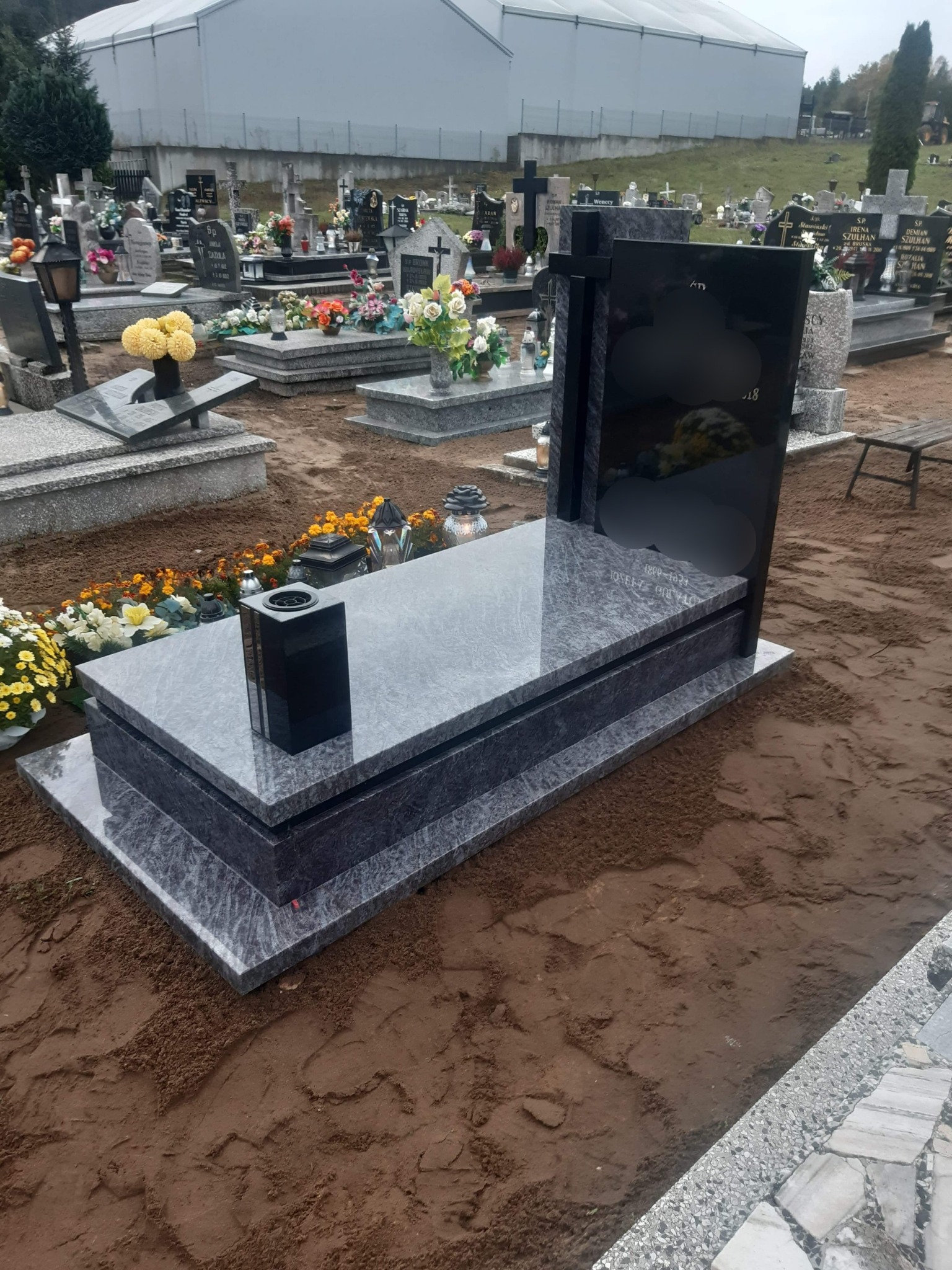 Nowy, szary nagrobek z czarną tablicą w otoczeniu innych grobów na cmentarzu, z kwiatami i zniczami.