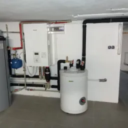 Instalacja kotła grzewczego Panasonic Aquarea z zbiornikiem buforowym i instalacją hydrauliczną w kotłowni, widoczne rury, zawory, filtry i osprzęt elektryczny.