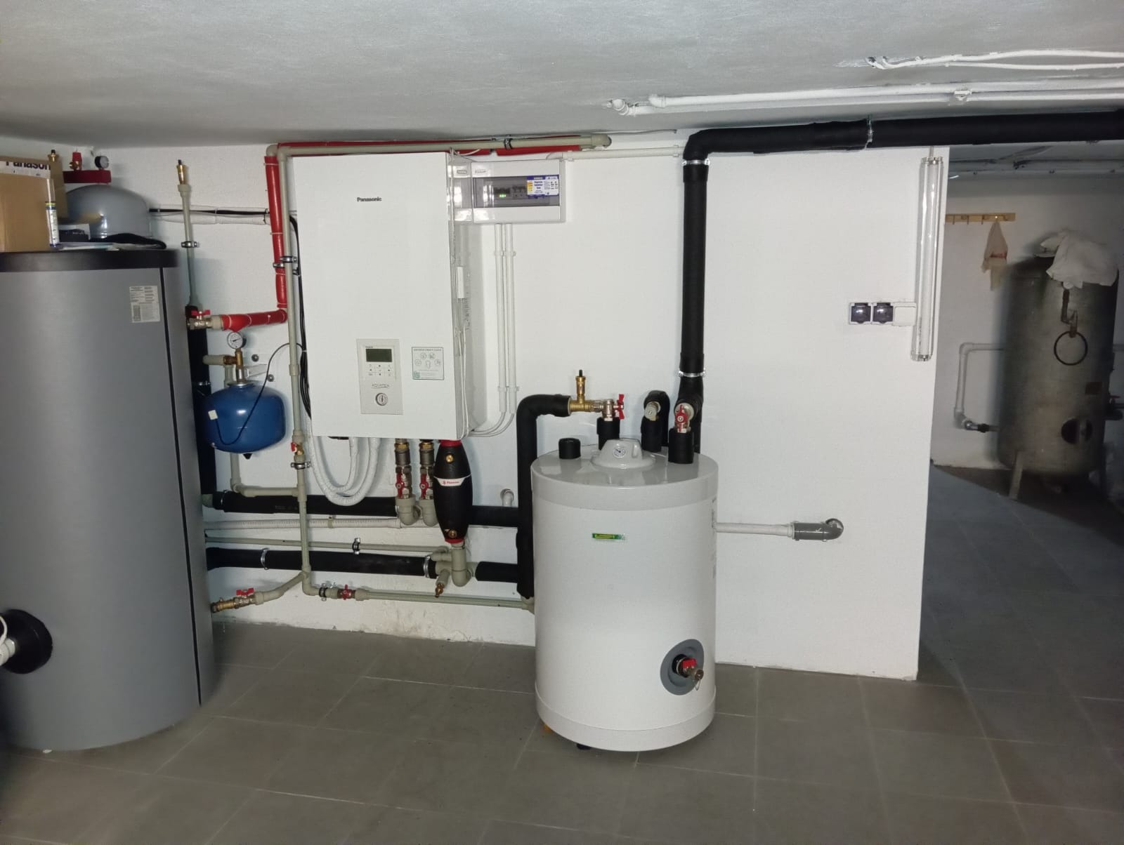 Instalacja kotła grzewczego Panasonic Aquarea z zbiornikiem buforowym i instalacją hydrauliczną w kotłowni, widoczne rury, zawory, filtry i osprzęt elektryczny.