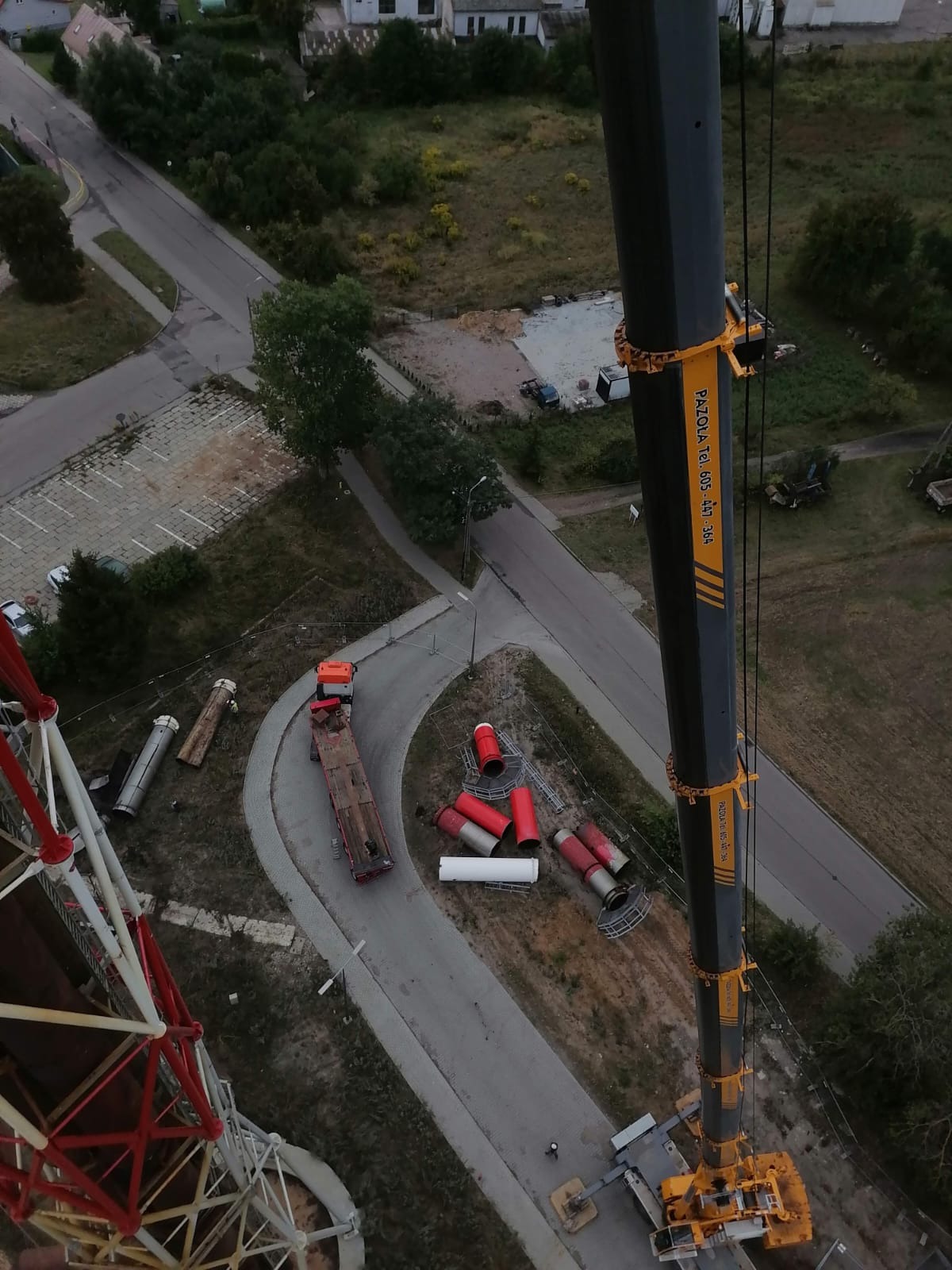 Widok z góry na żuraw wieżowy podczas montażu segmentów wieży, widoczna ciężarówka z platformą i elementy konstrukcyjne na ziemi.