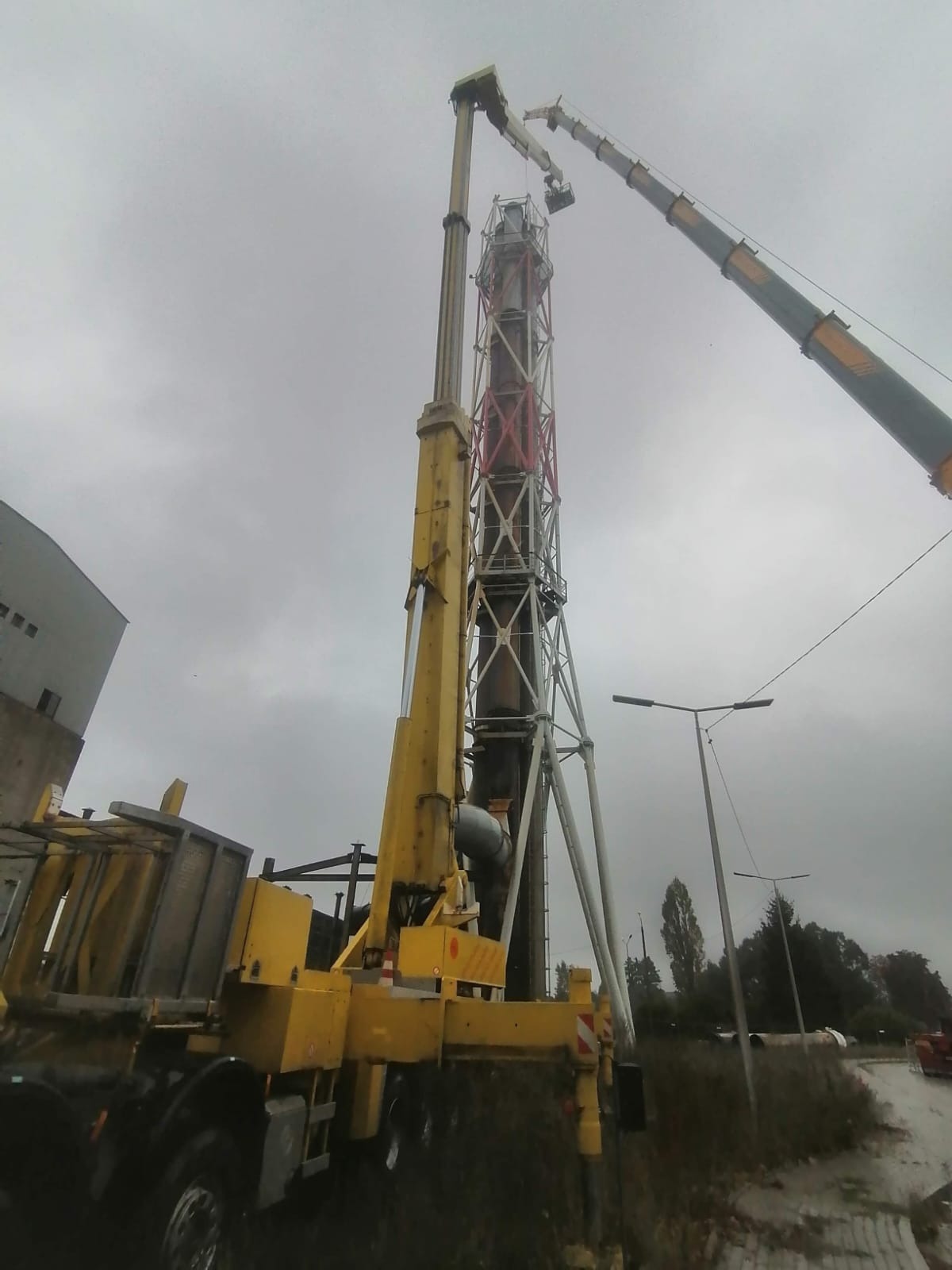 Dwa żółte dźwigi teleskopowe pracują przy demontażu wysokiego, metalowego komina z elementami pomalowanymi na czerwono i biało, na tle pochmurnego nieba.