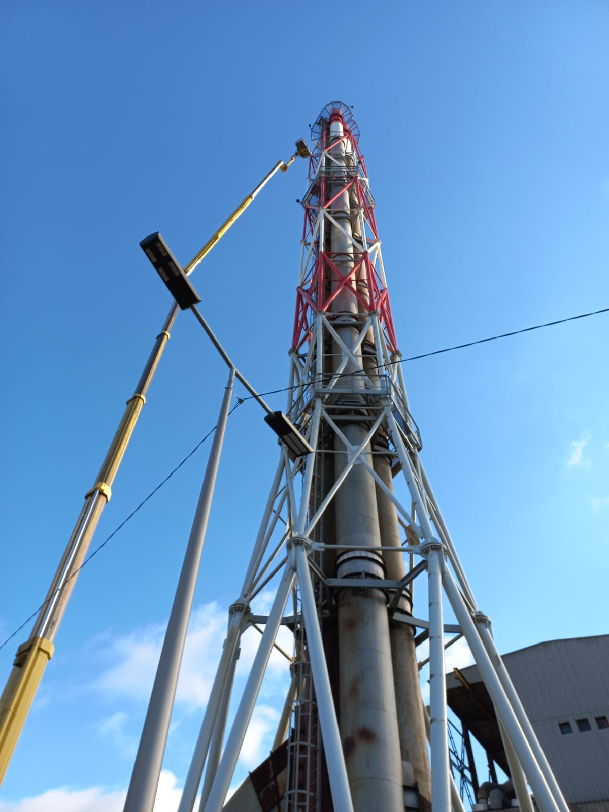 Wysoki, stalowy komin przemysłowy z czerwoną i białą kratownicą, widziany z dołu na tle błękitnego nieba, z wysięgnikiem żółtego dźwigu w trakcie prac konserwacyjnych lub naprawczych na wysokości.