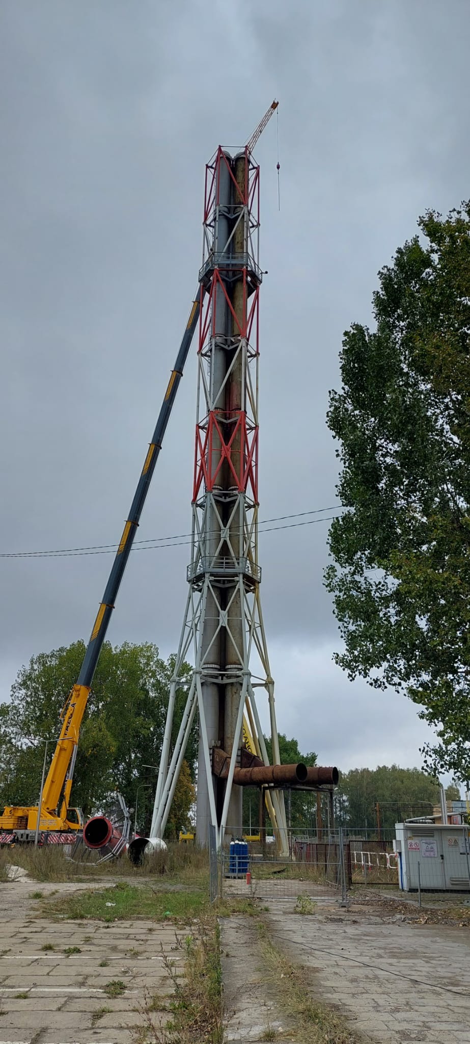 Demontaż wysokiego komina przemysłowego z użyciem dźwigu, widoczne elementy konstrukcji stalowej i rurociągi.