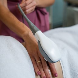 Salveo-Bioesthetic - Urządzenie do modelowania sylwetki w kontakcie ze skórą pacjenta, dłoń w złotym pierścionku, biały ręcznik w tle, zabieg na ciało w gabinecie.