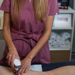 Salveo-Bioesthetic - Osoba w różowym uniformie wykonuje zabieg na nodze pacjenta przy użyciu specjalistycznego urządzenia. W tle widoczny regał z ręcznikami i materiały informacyjne.