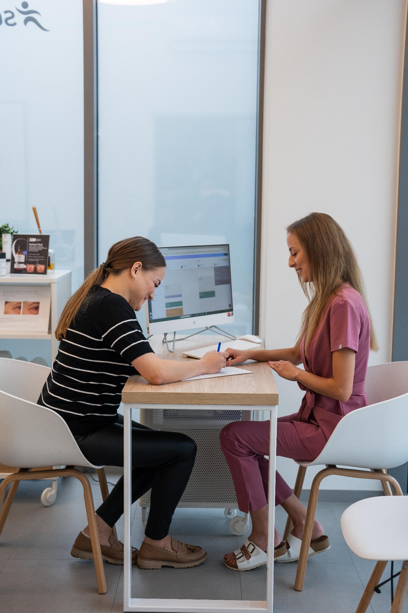 Konsultacja w gabinecie: kobieta w stroju medycznym omawia plan zabiegów z klientką, która wypełnia formularz. Widoczny monitor z kalendarzem wizyt.