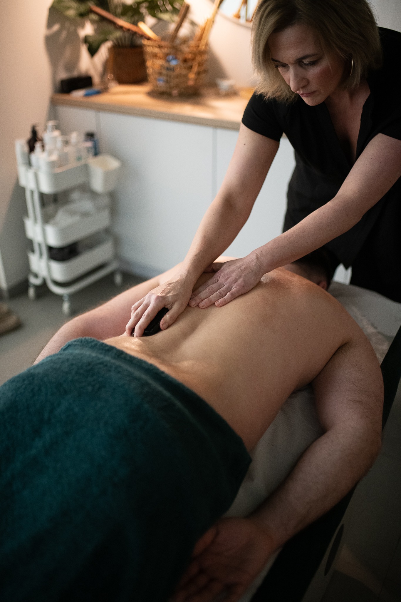 Masażysta wykonuje masaż kamieniami na plecach klienta leżącego na stole do masażu, w tle widać białe meble i akcesoria do masażu.