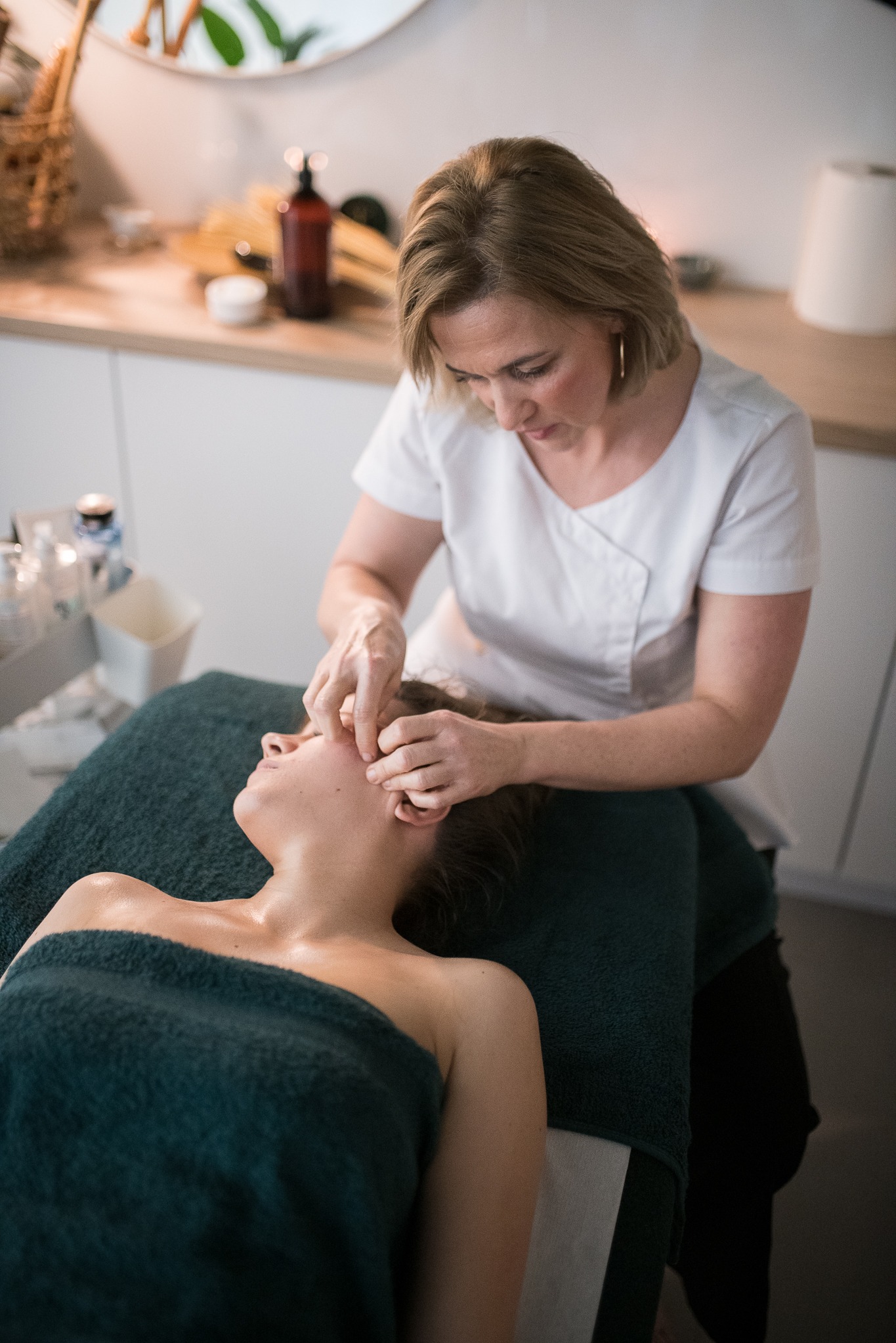 Kobieta w białym uniformie wykonuje delikatny masaż twarzy klientki leżącej na zielonym ręczniku w gabinecie. Widoczne kosmetyki i akcesoria.