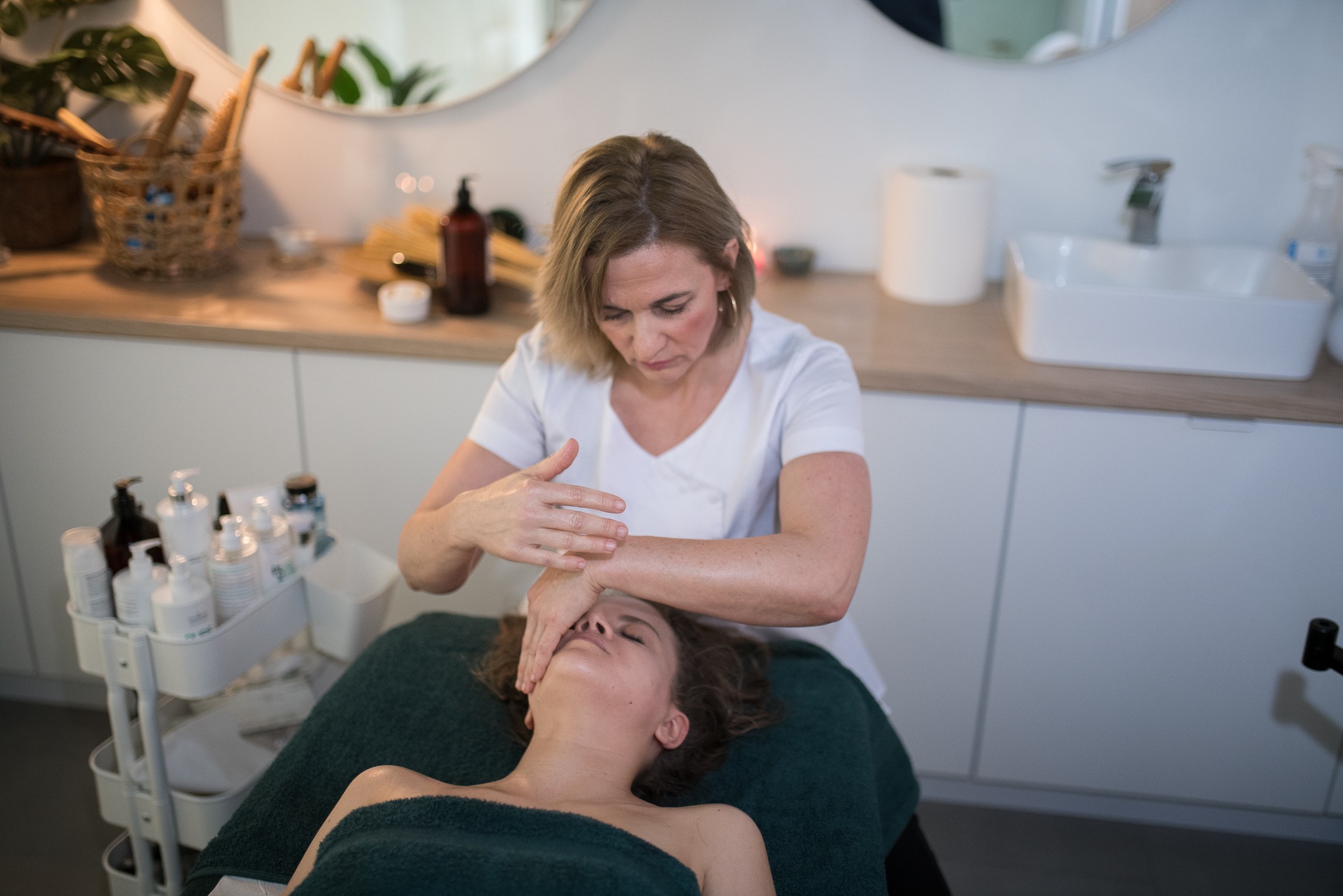 Kobieta wykonuje masaż twarzy klientce leżącej na zielonym ręczniku. W tle widać kosmetyki i akcesoria w jasnym wnętrzu gabinetu.